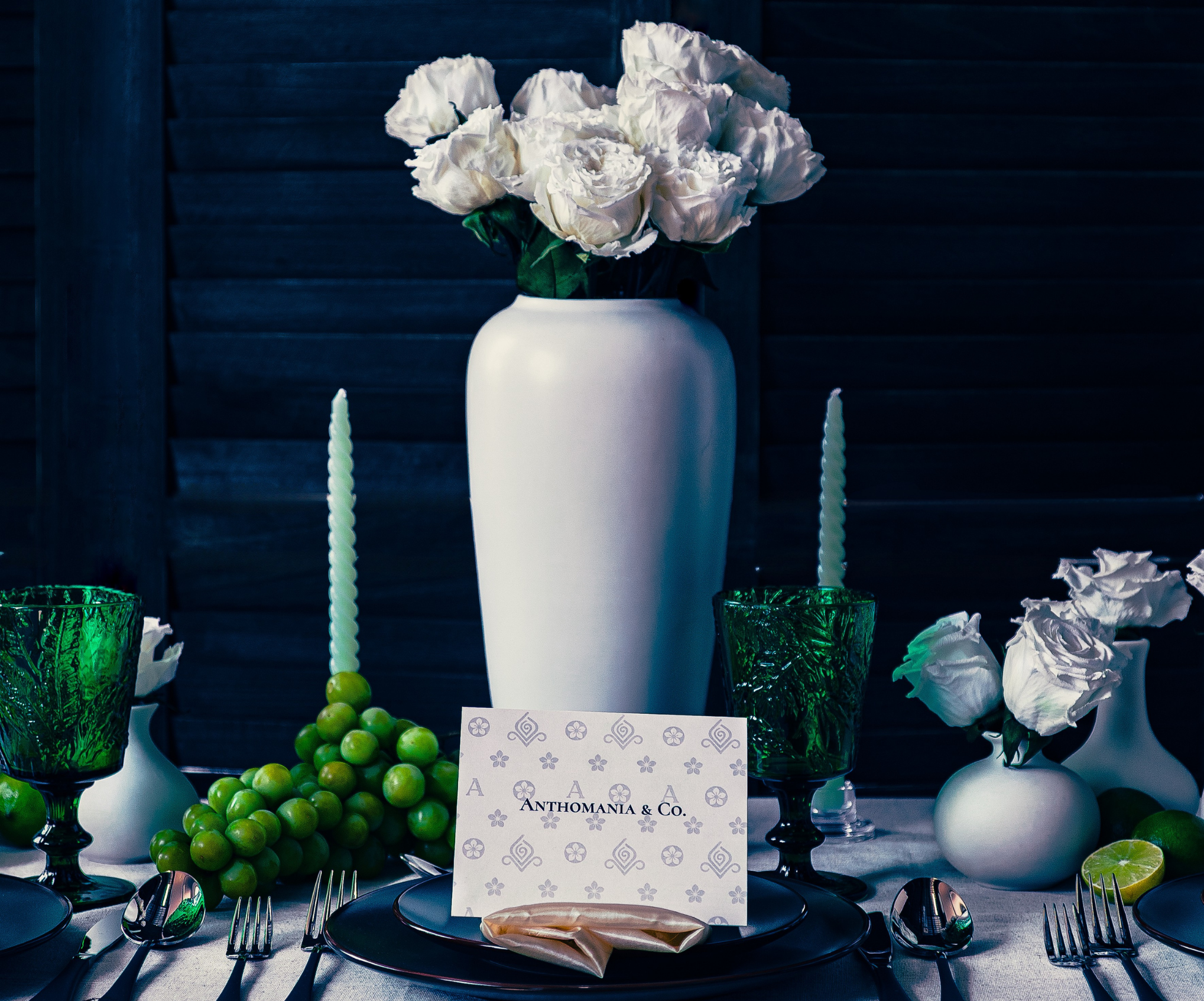 Elegant table setting with white roses in a vase, green grapes, and decorative cards on a dark background.